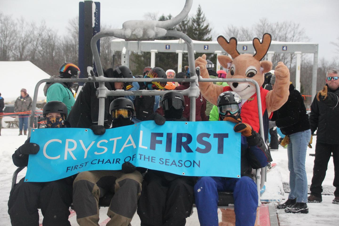 Skiers on a lift with a Buck mascot and a blue banner in snowy surroundings.
