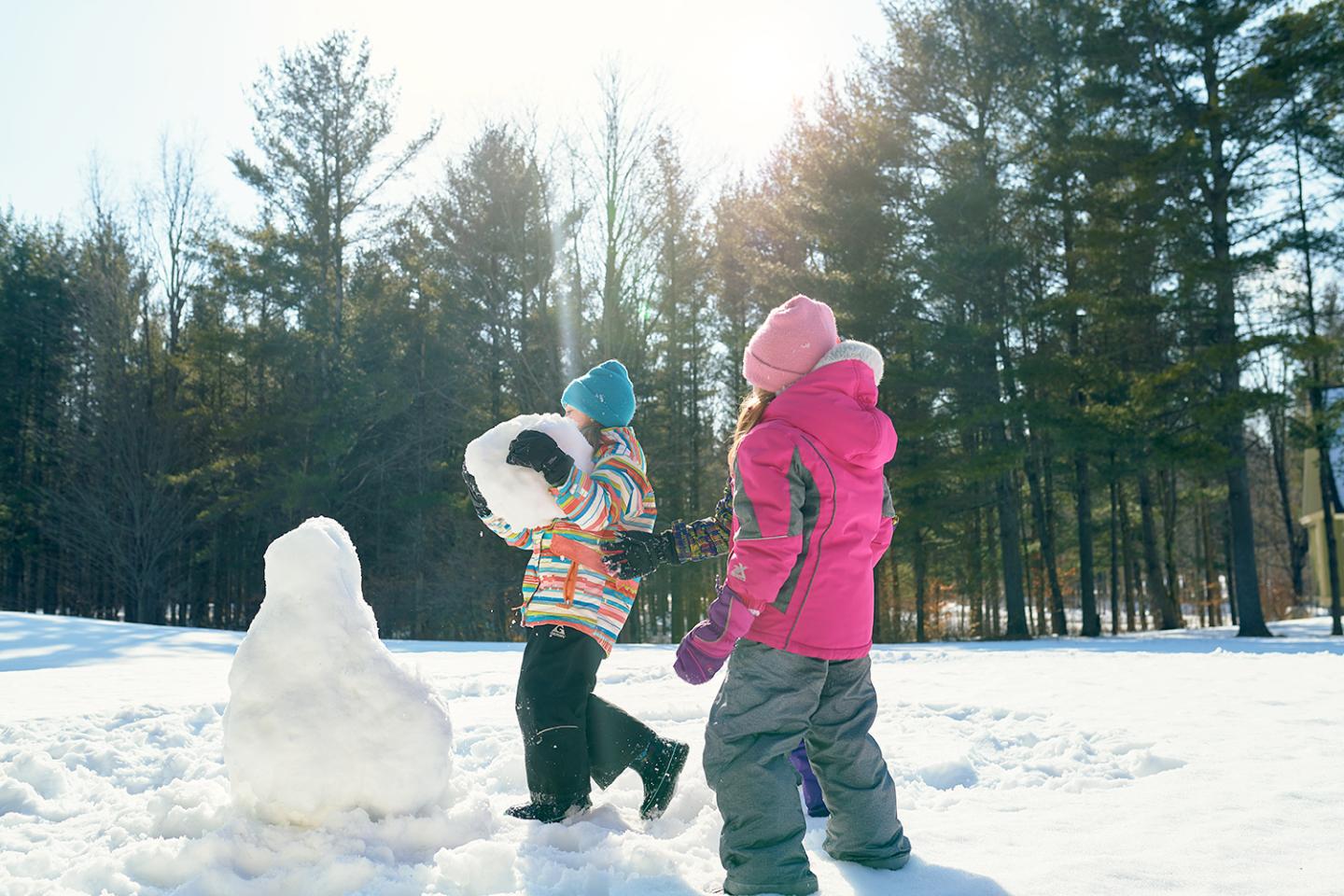 Children in colorful winter clothes building a snowman in a snowy forest.