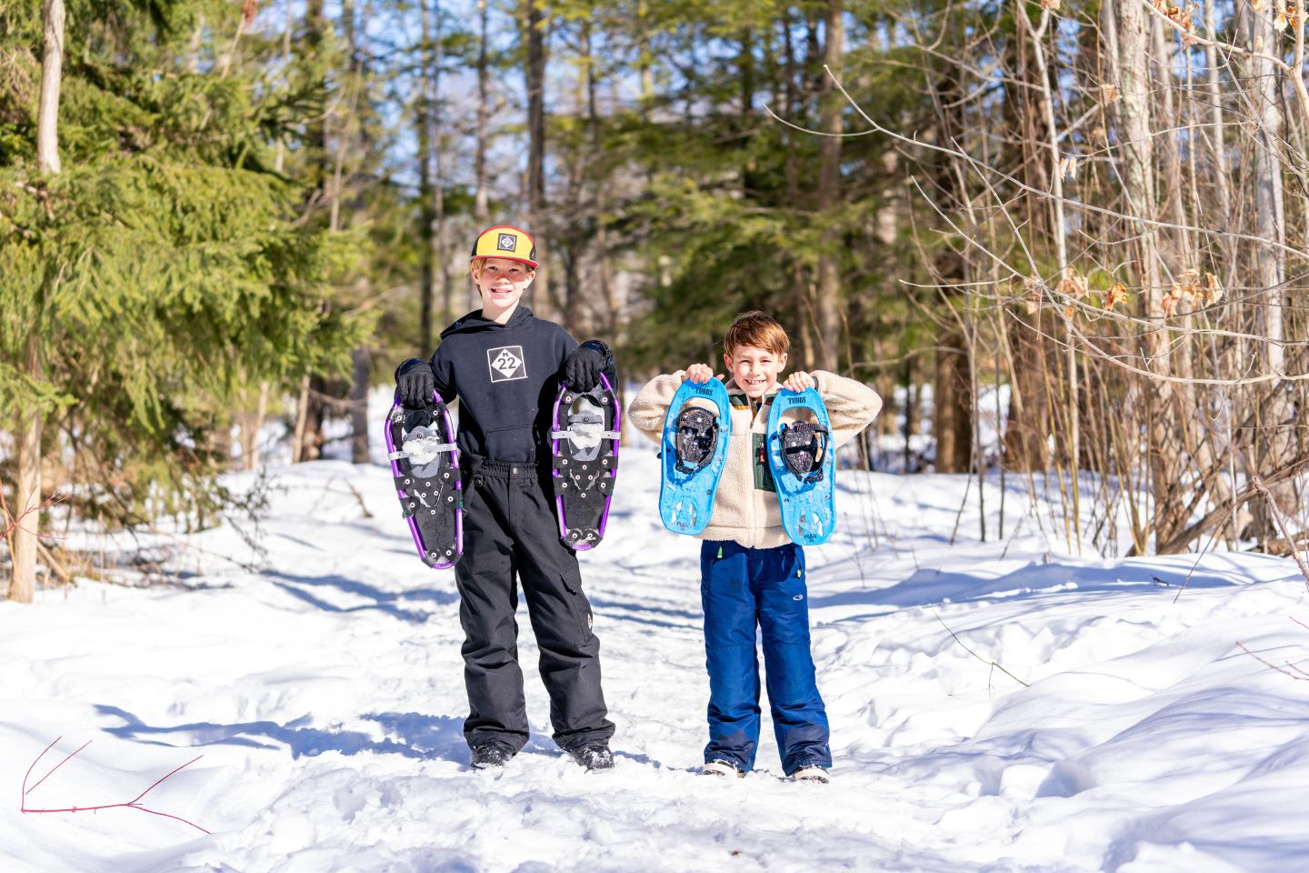 Two children holding snowshoes, standing on a snowy forest trail.