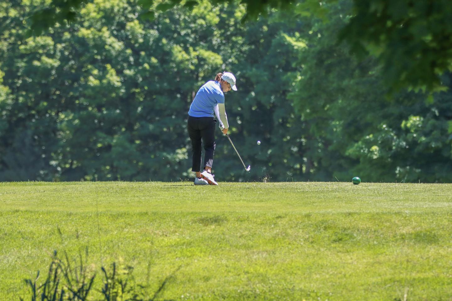 Female golfer swinging on a sunny green course with trees in the background.