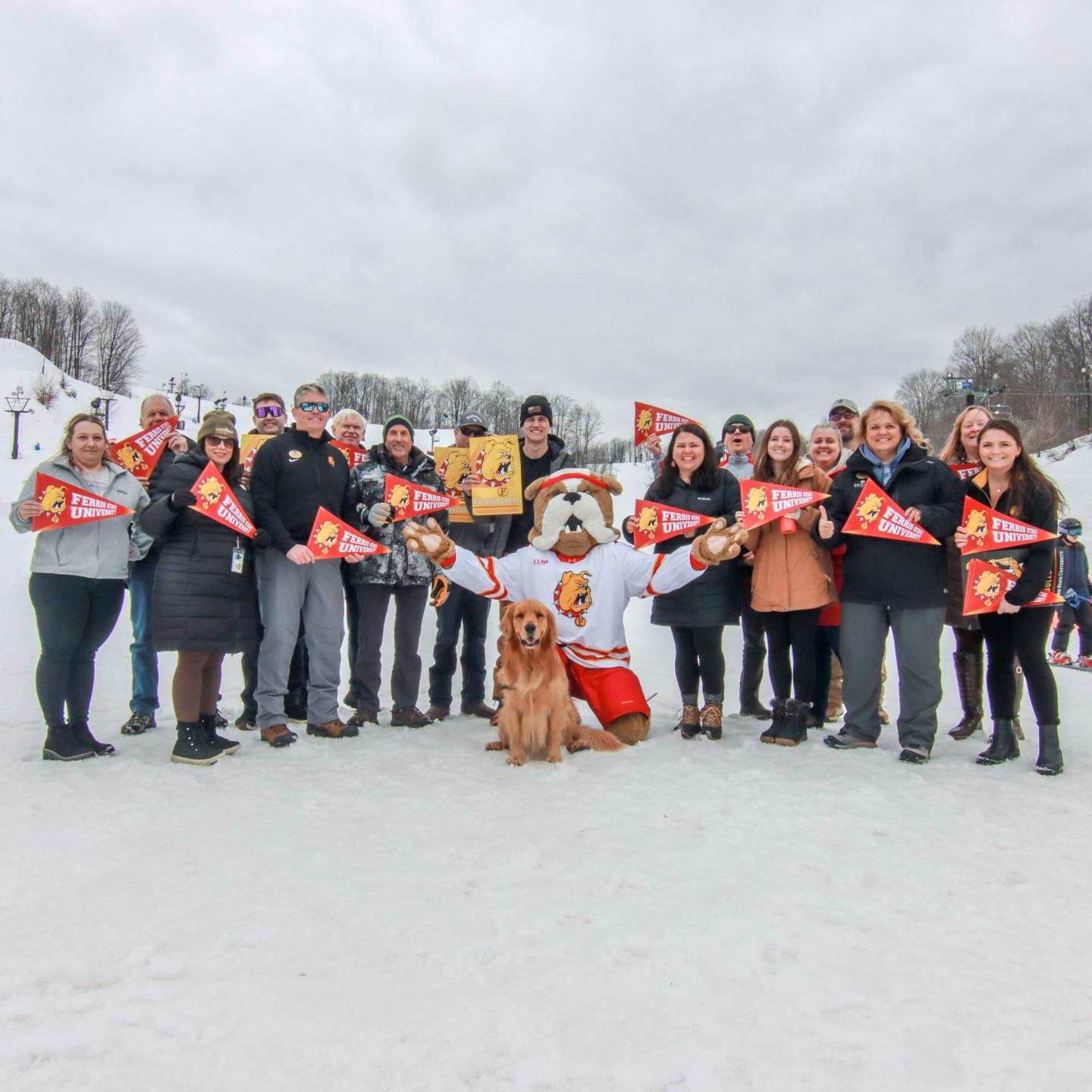 Group of people with mascot and dog on snowy ground, holding red pennants, cloudy sky.
