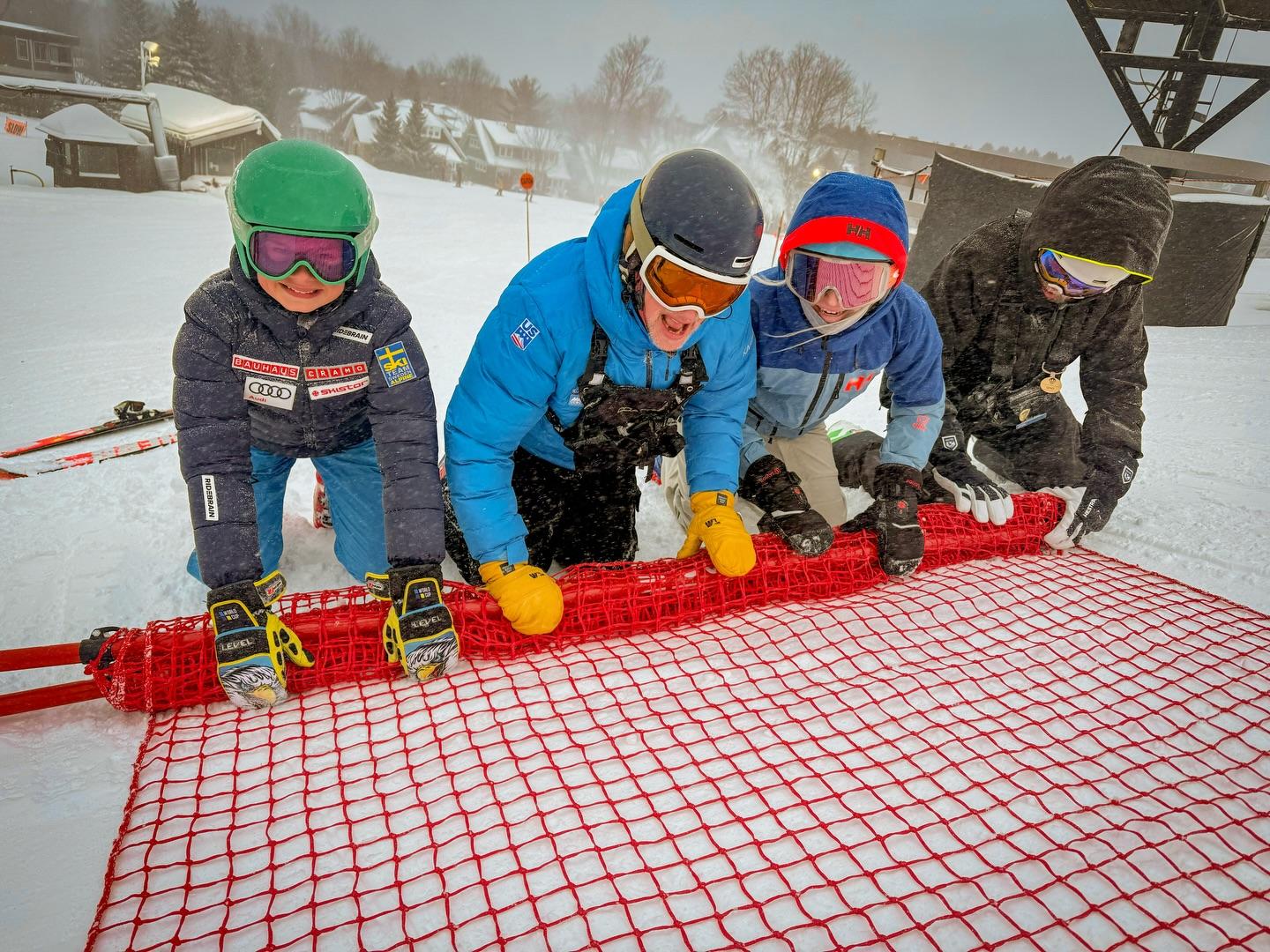 Four people in winter gear smiling and rolling a red snow fence on a snowy day.