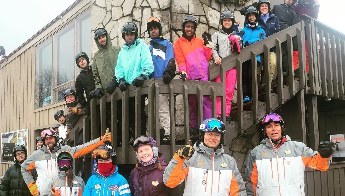 Group of skiers wearing winter gear, smiling on wooden stairs and outside building.