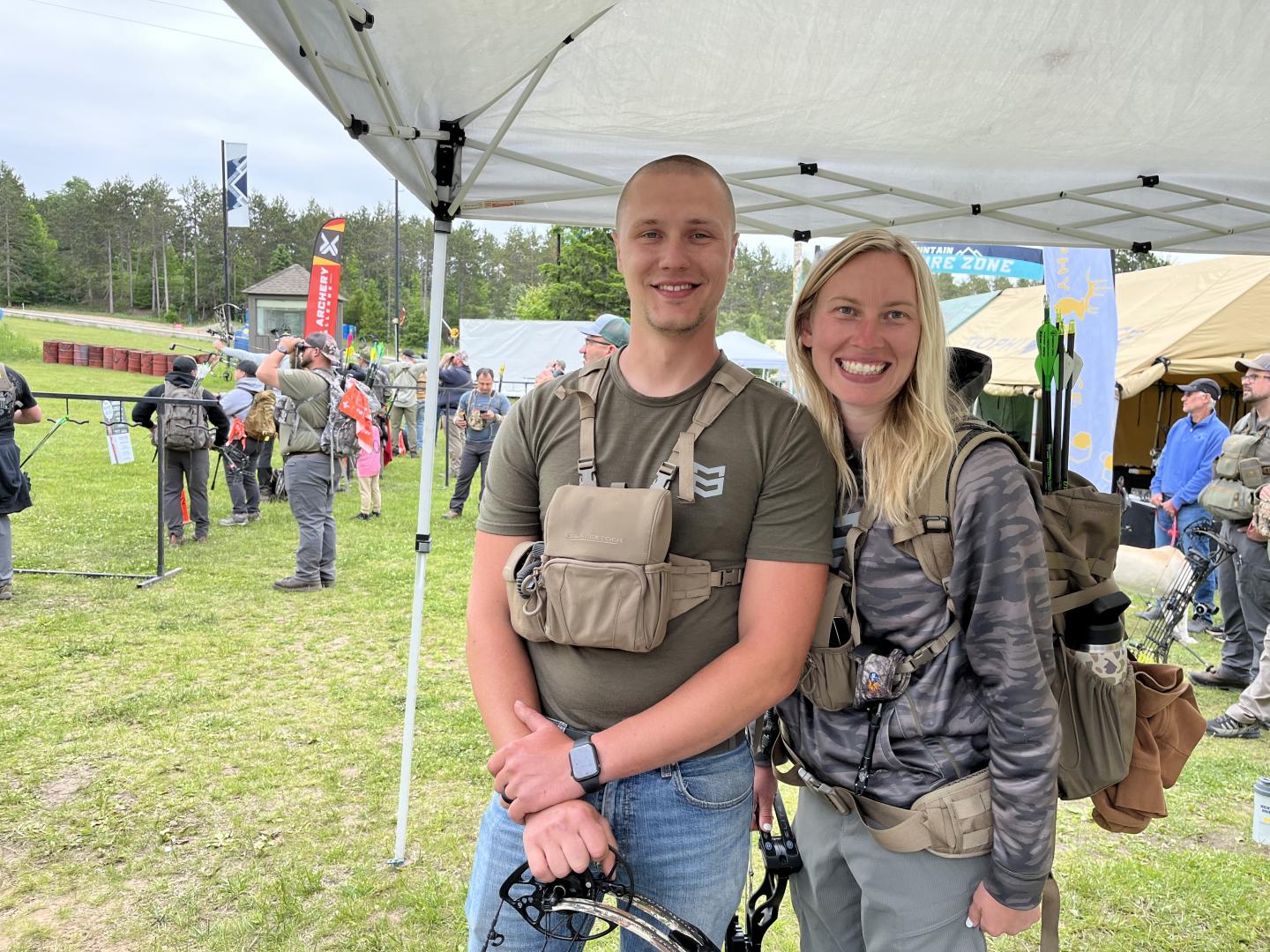 Smiling couple under a canopy at an outdoor event, with people and tents in the background.