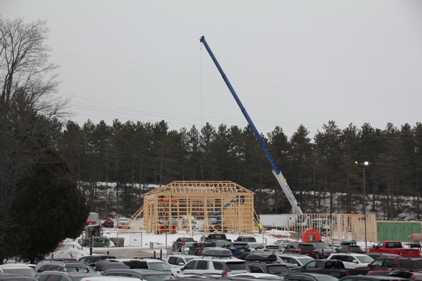 Construction site with crane and wooden framework, surrounded by parked cars in snowy setting.