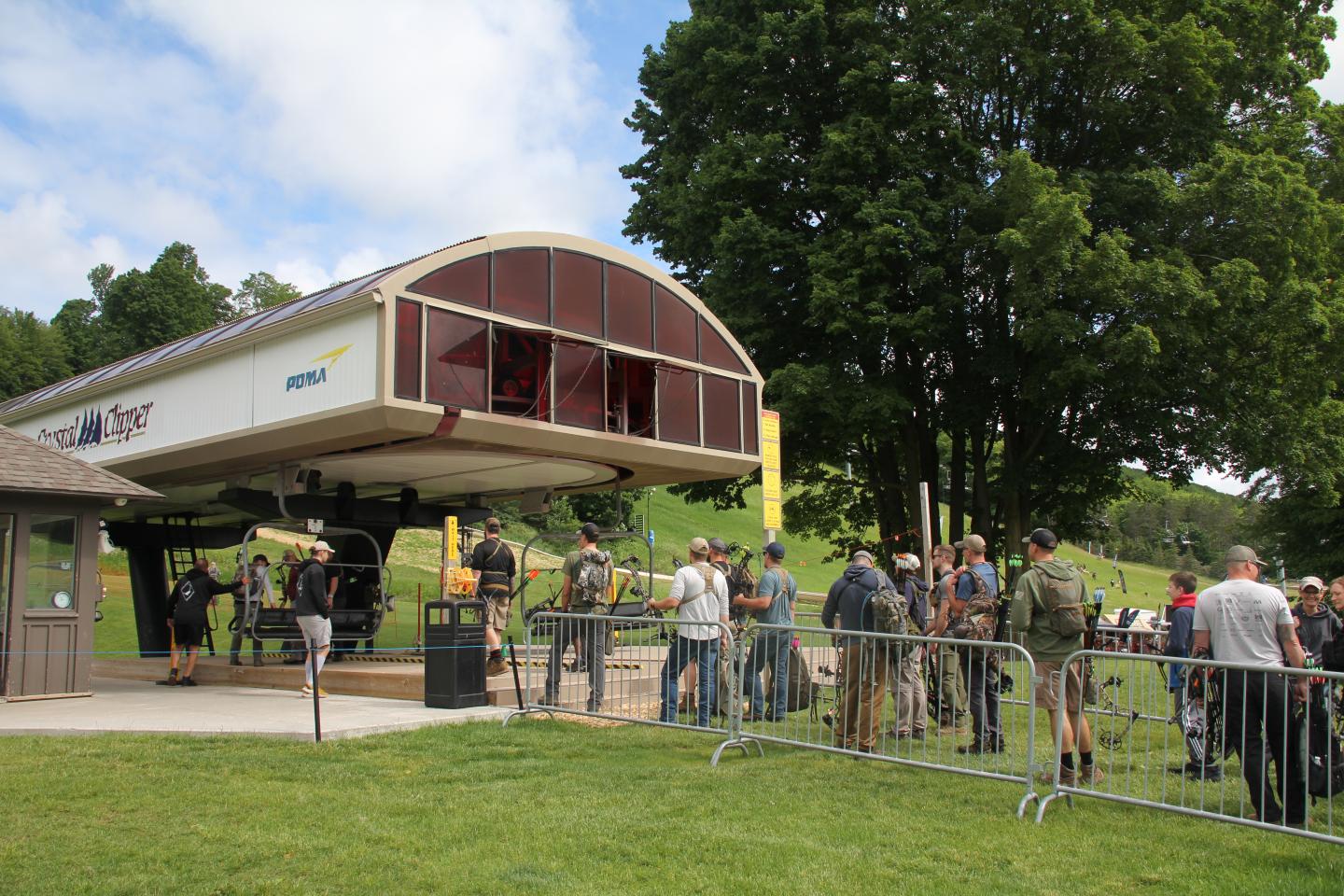 Ski lift with people in line, surrounded by trees and a cloudy sky.