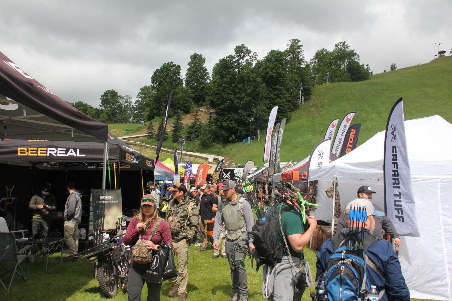 Crowd at an outdoor gear expo with tents and flags, set on a grassy hill.