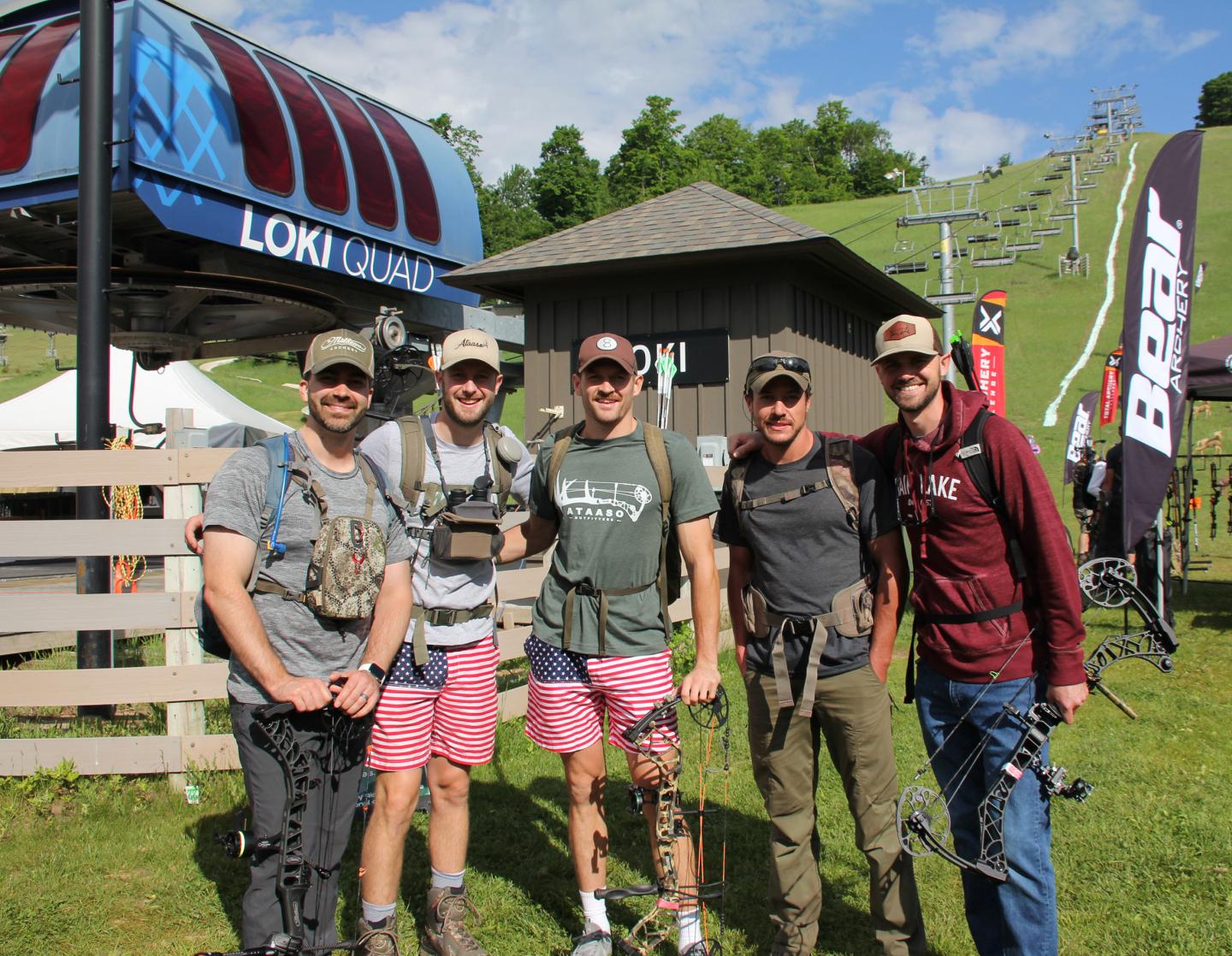 Five men outdoors with bows, smiling under a blue sky near ski lift and flags.