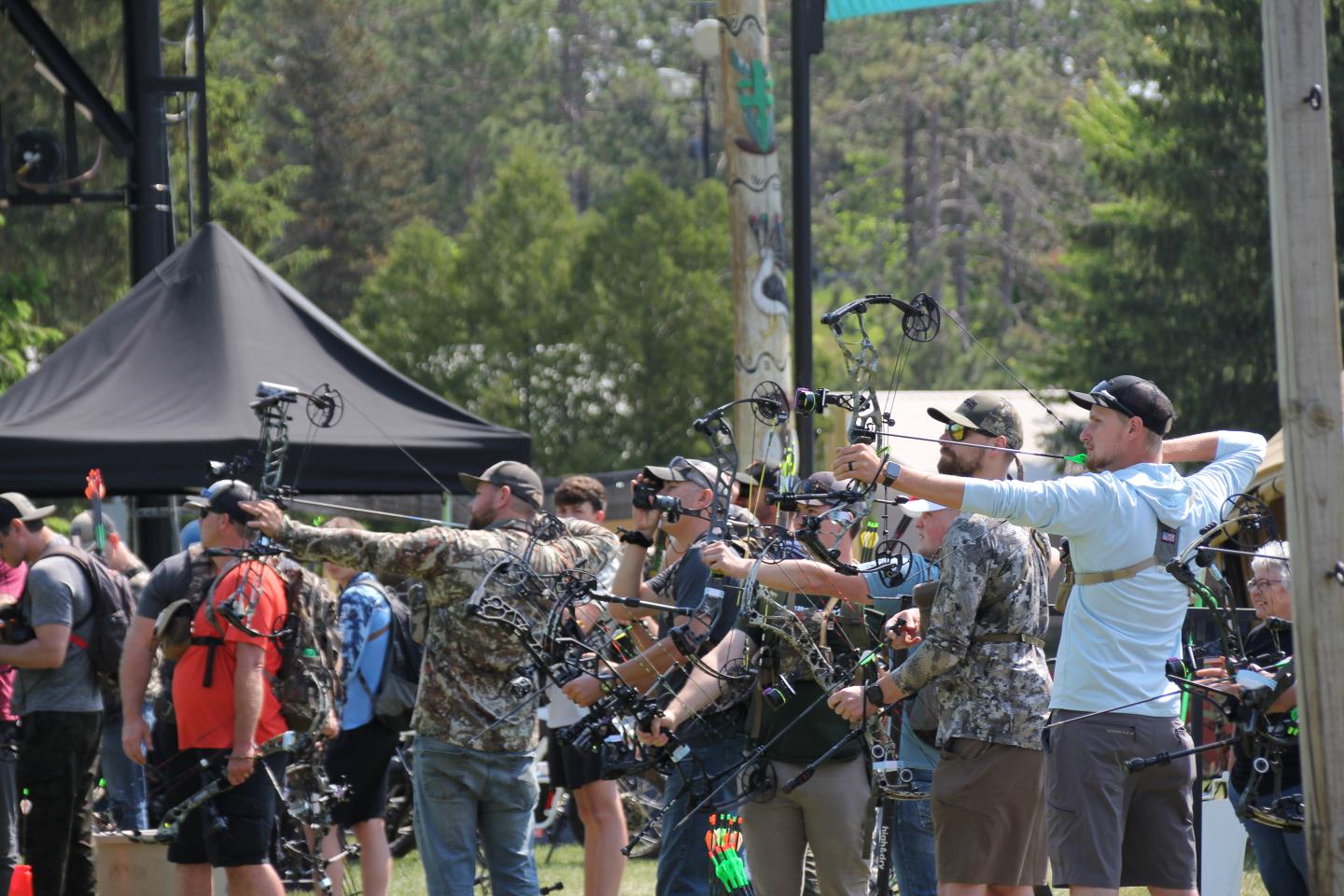 Archers aim compound bows at an outdoor event, surrounded by trees and tents.