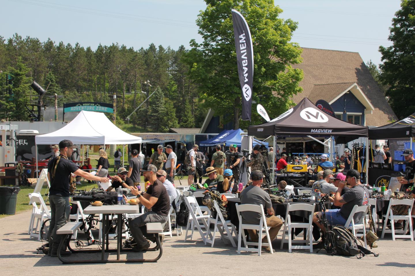 Outdoor gathering with people sitting at tables, surrounded by tents and trees.