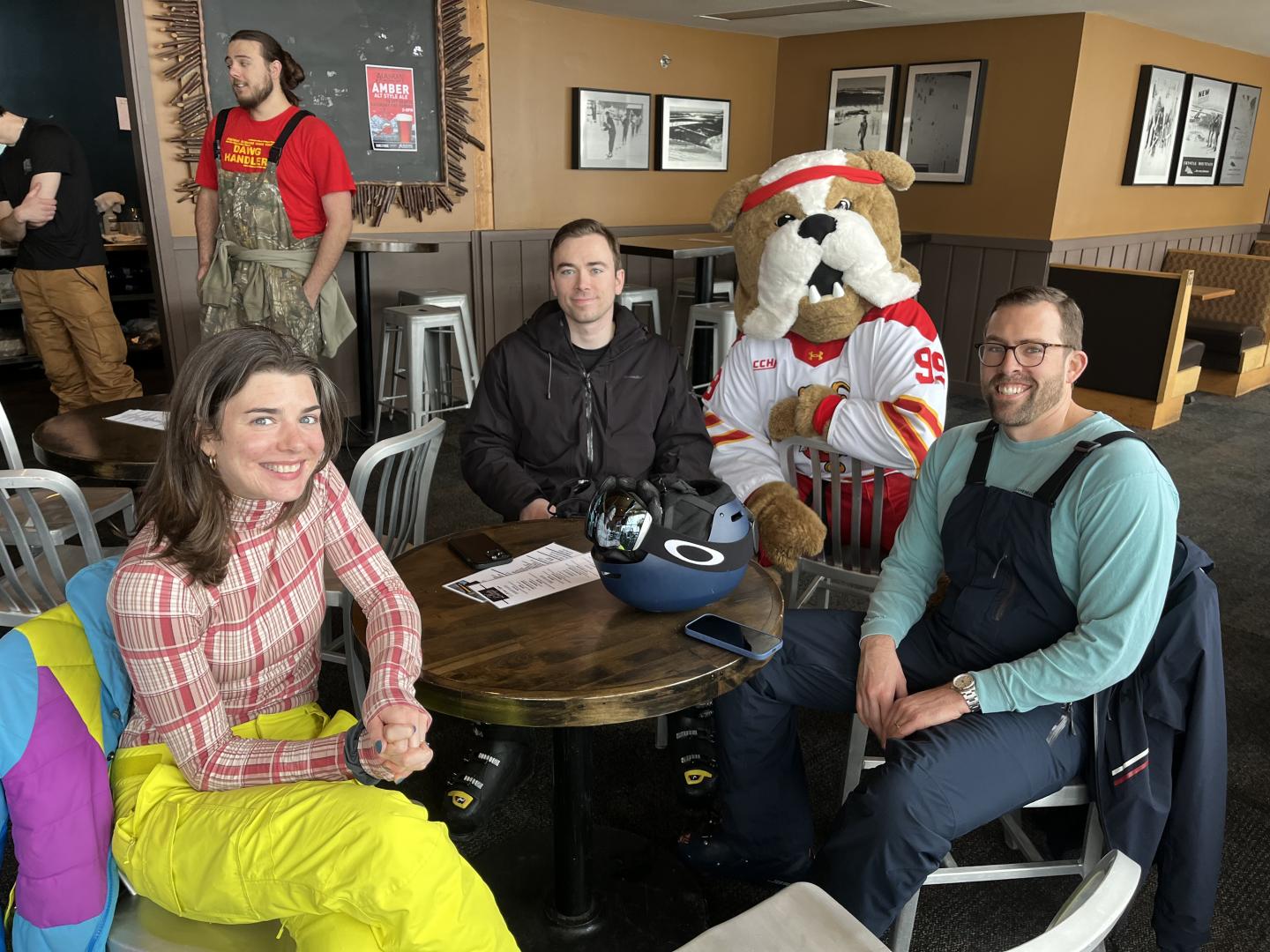 Four people and a mascot in ski gear at a café table, smiling.