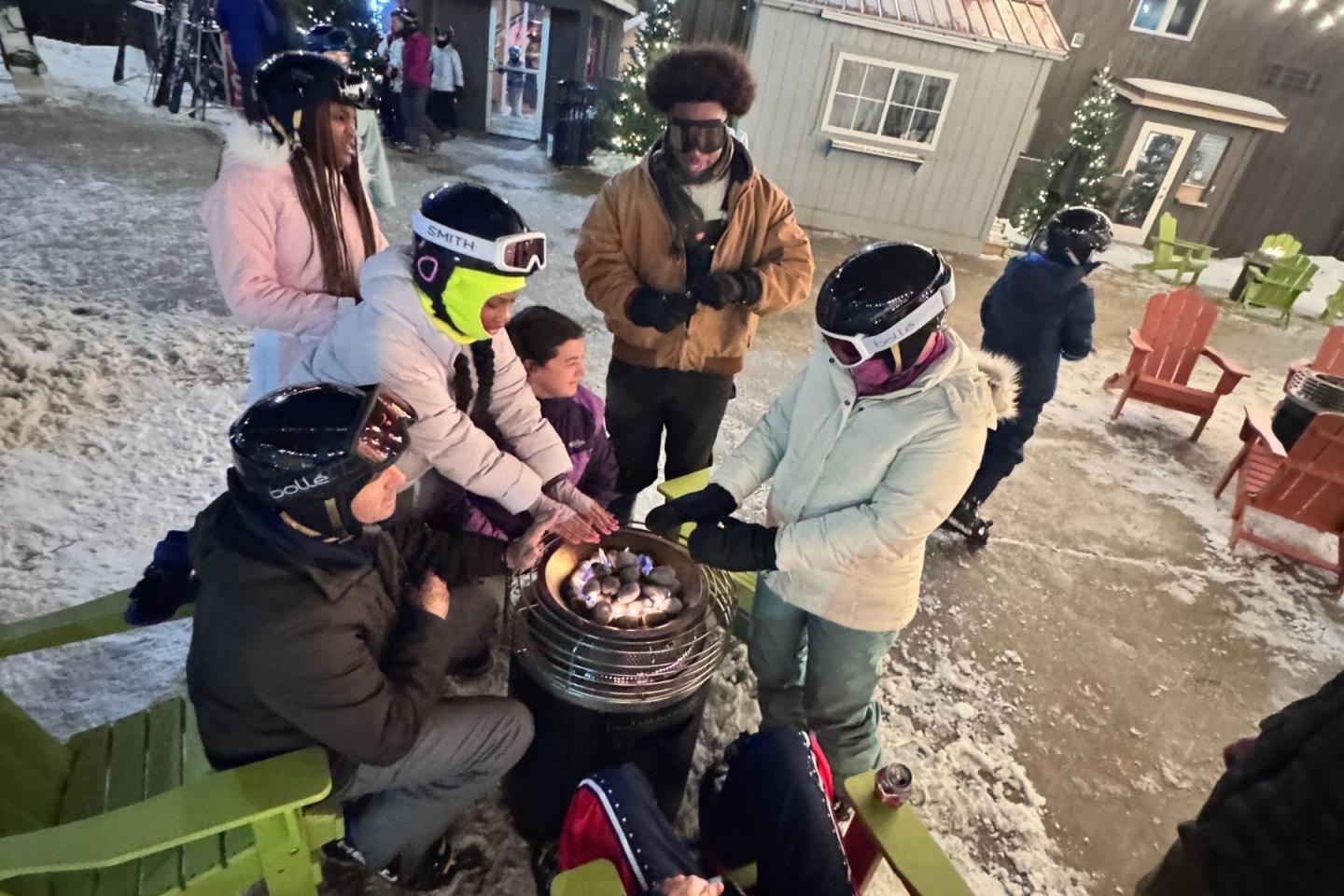 People in winter gear gather around a small fire pit in a snowy outdoor setting.