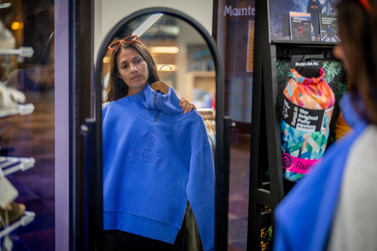 Woman holding a blue sweater in front of a mirror in a store.