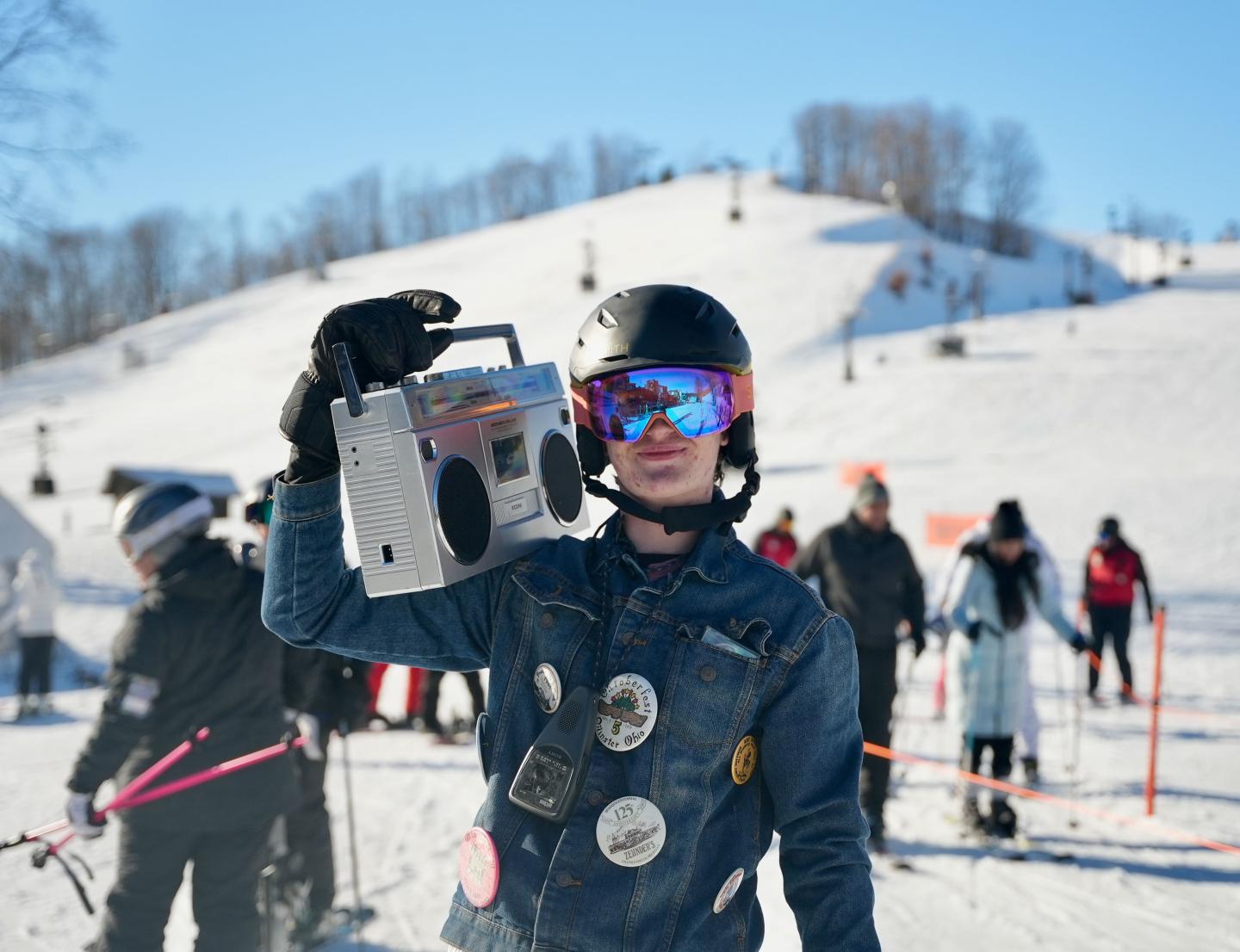 Snowboarder holding a boombox, wearing a helmet and goggles on a snowy hill.