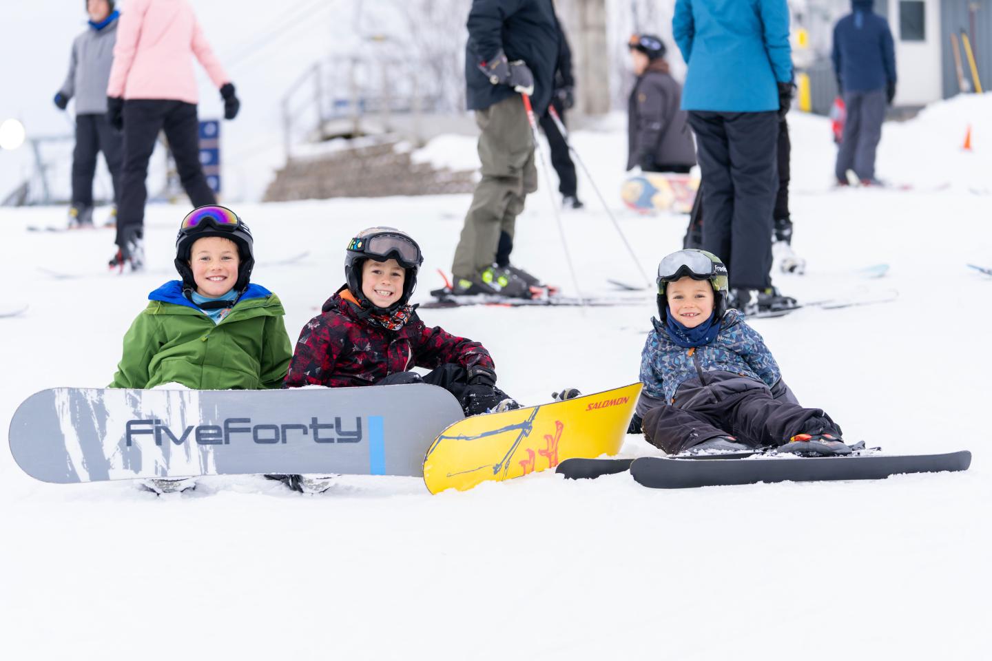 Kids in snow gear sitting with snowboards on a snowy slope.