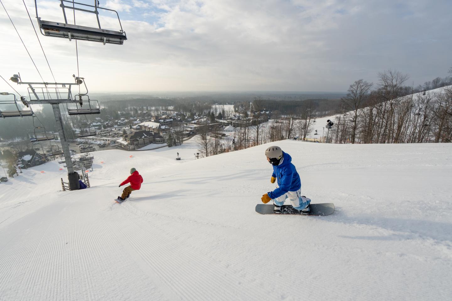 Snowboarders on a snowy slope, with ski lift and town below.