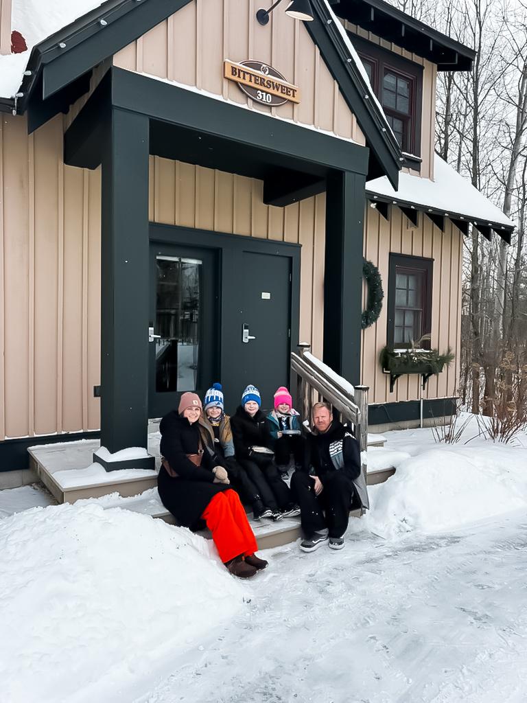 People sitting on snowy steps in front of a cottage, wearing winter clothes.