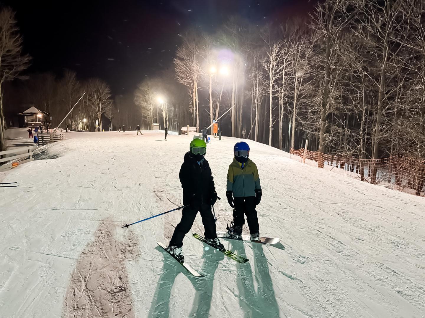 A skier and snowboarder on a lit snowy slope at night, surrounded by trees.