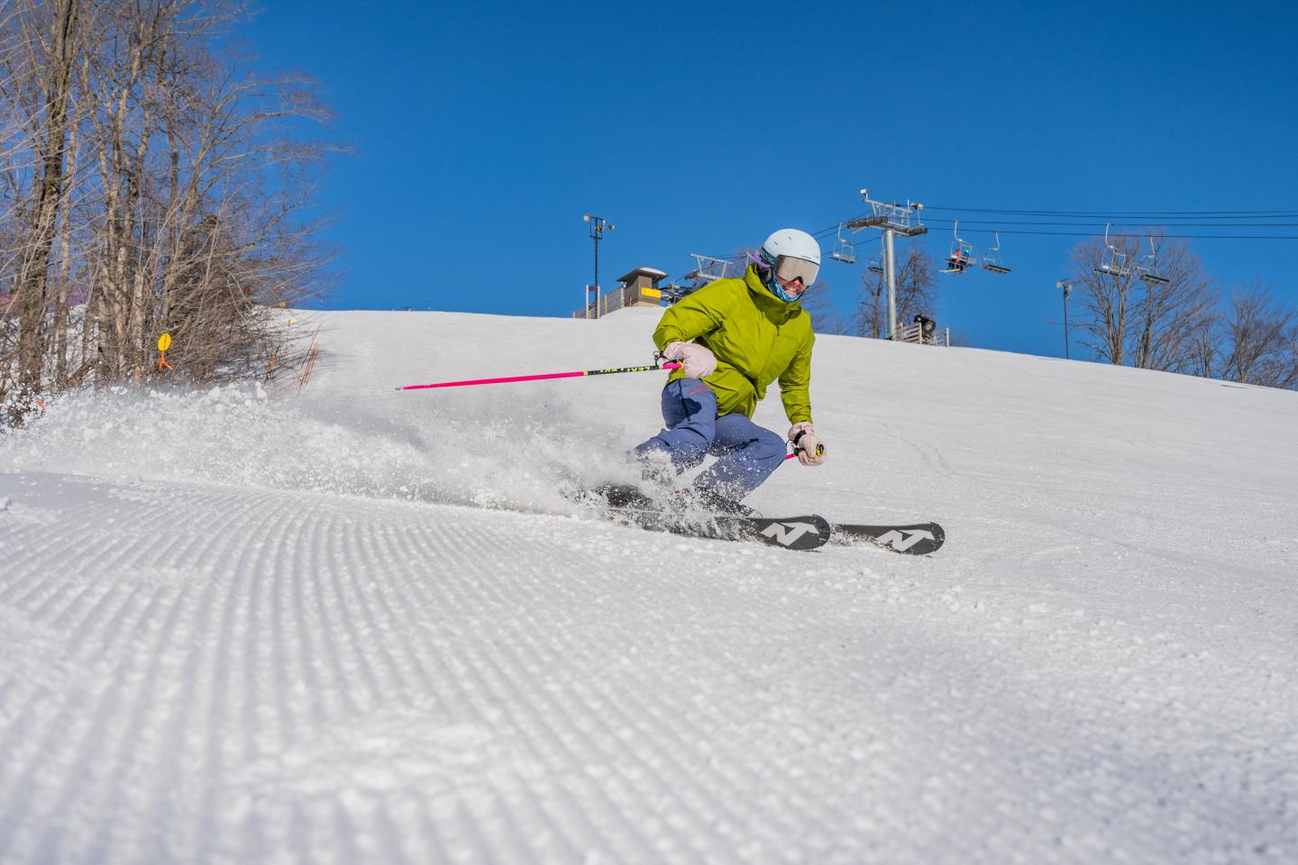 Skier in green jacket descending snowy slope under clear blue sky.