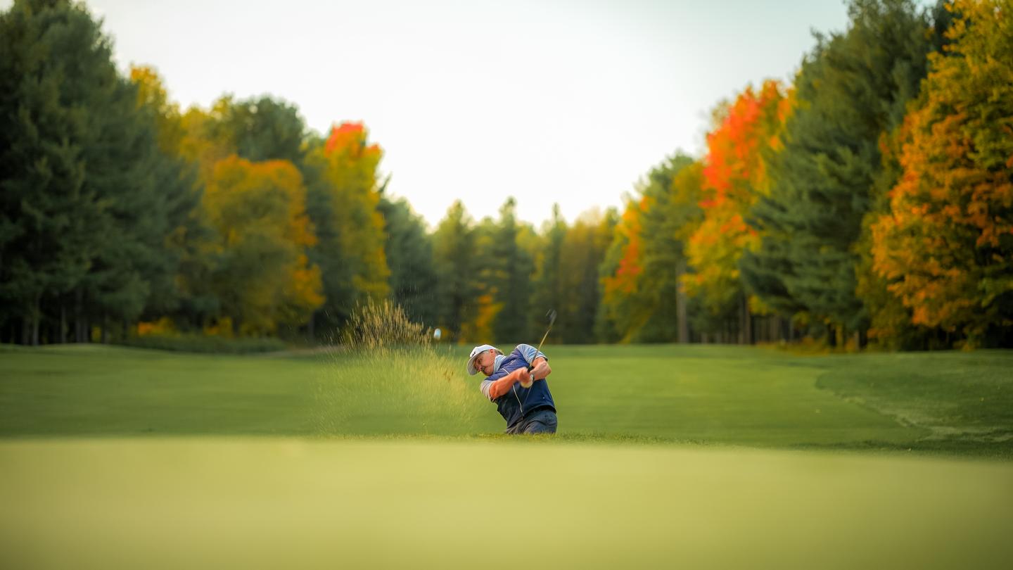 Golfer hitting from a sand bunker, surrounded by autumn trees.