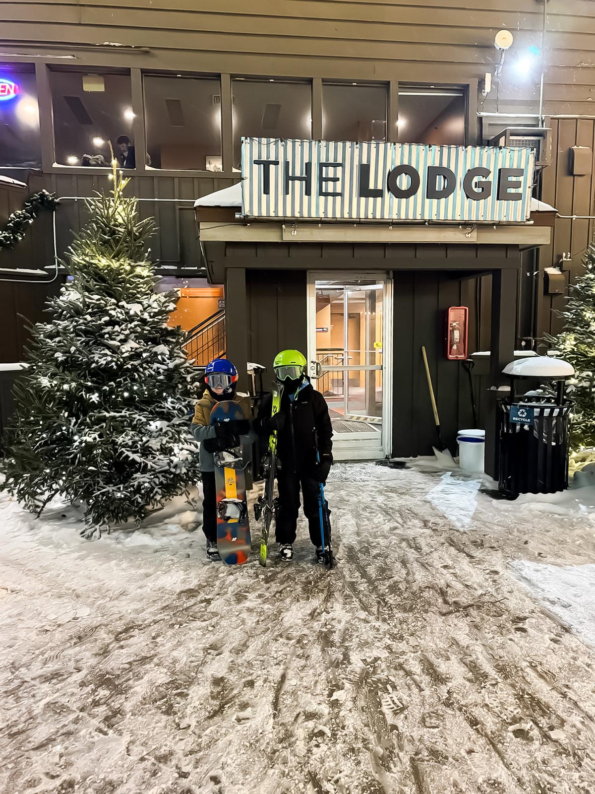 Two people in ski gear outside a snowy lodge entrance at night.