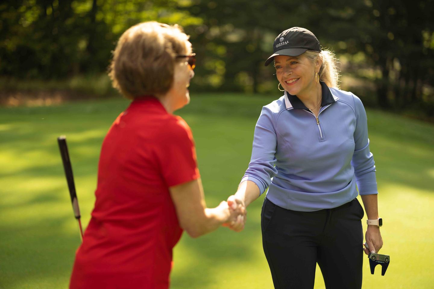 Two women smiling and shaking hands on a sunny golf course.