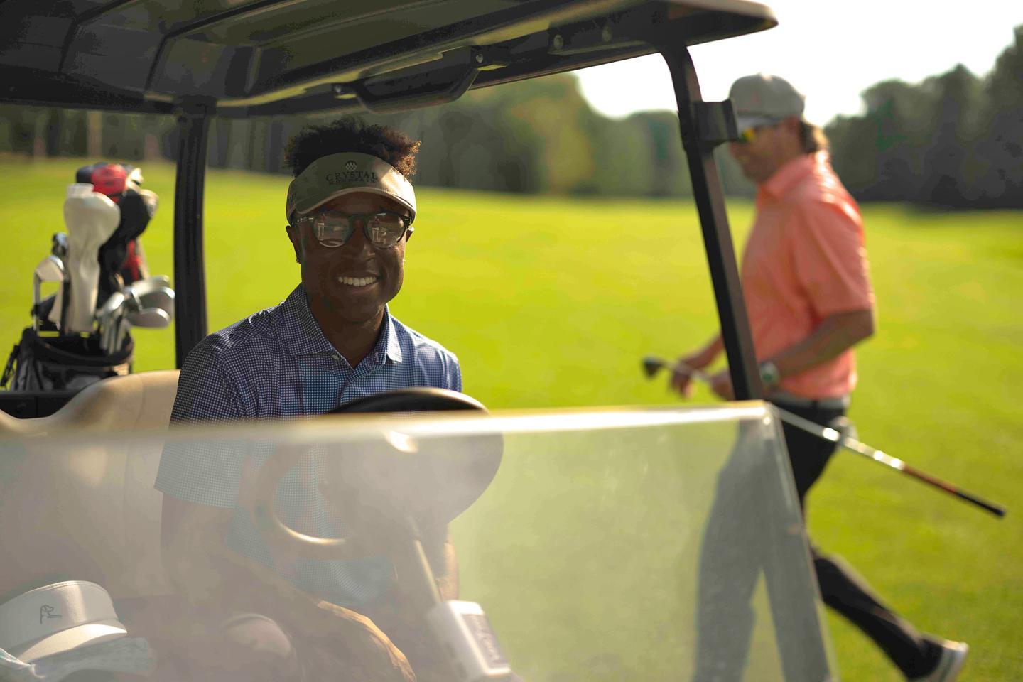 Golfers on a sunny day, one in a cart, another walking with clubs.