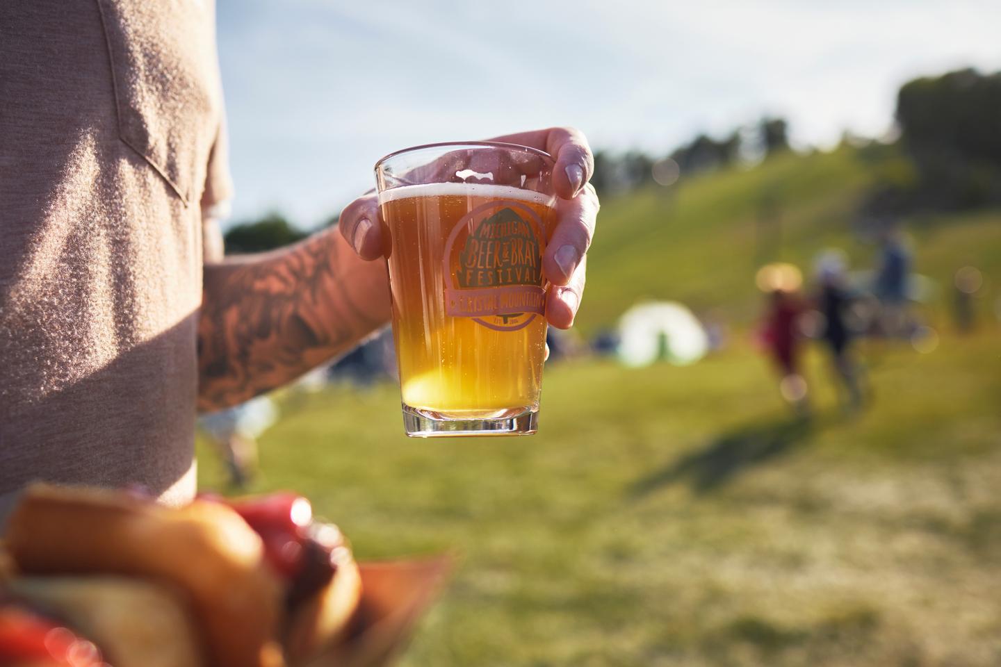 Glass of beer held outdoors at a sunny picnic with blurry people in background.