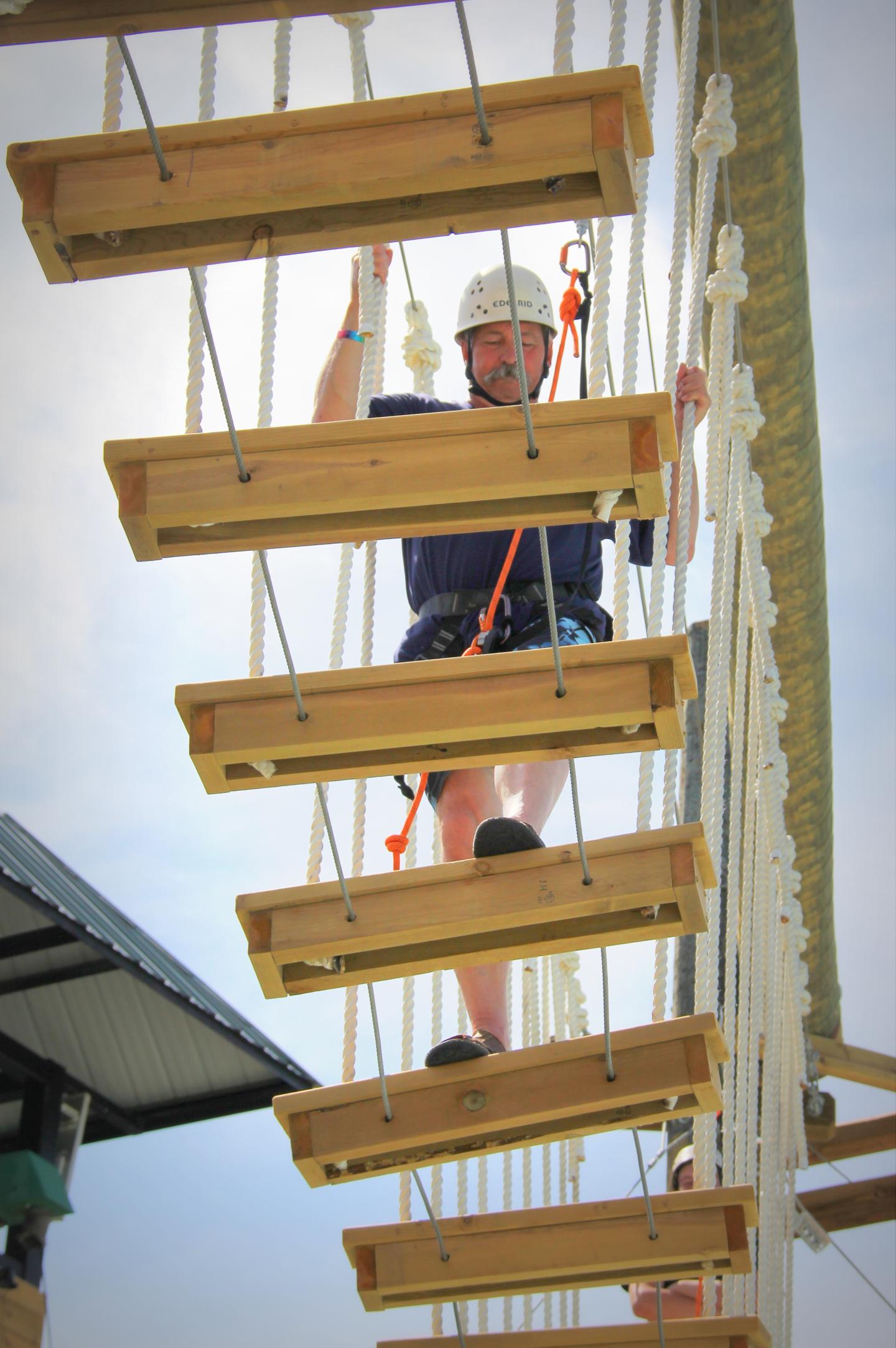 Person climbing a rope bridge, wearing a helmet and harness, sunny day.