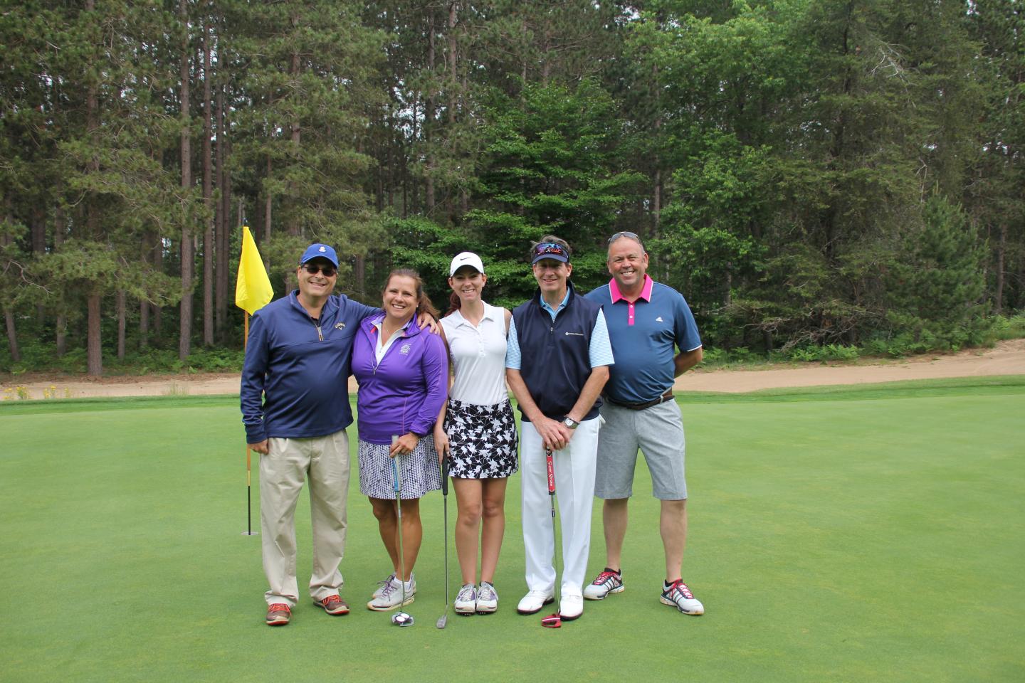 Five people posing together on a golf course green, with trees in the background.
