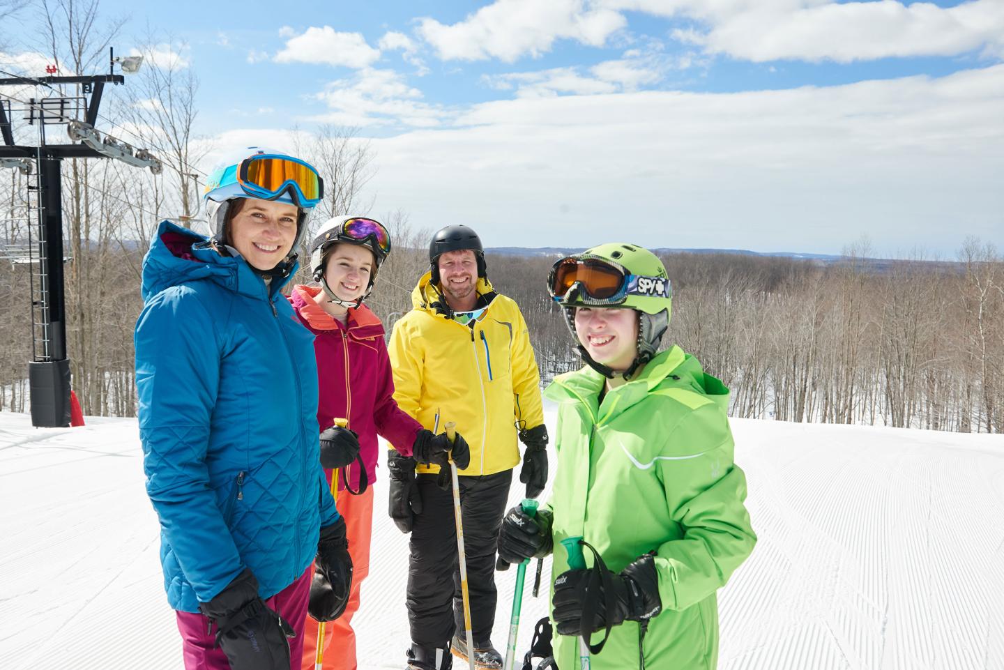 Four people in colorful ski gear stand smiling on a snowy slope under a blue sky.