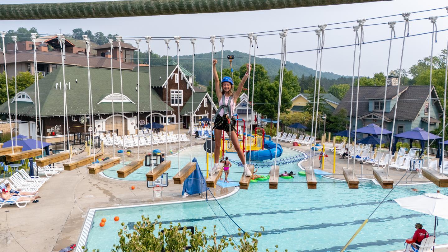 Child on a rope course above a swimming pool with scenic background.
