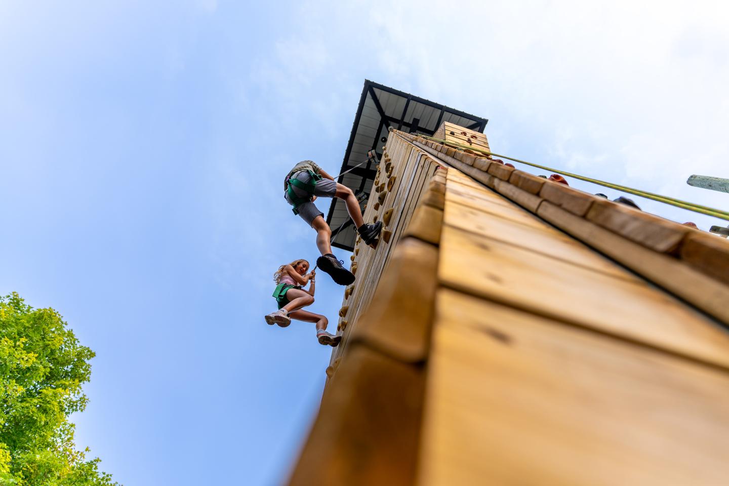 Two people climbing a tall wooden wall with a blue sky in the background.