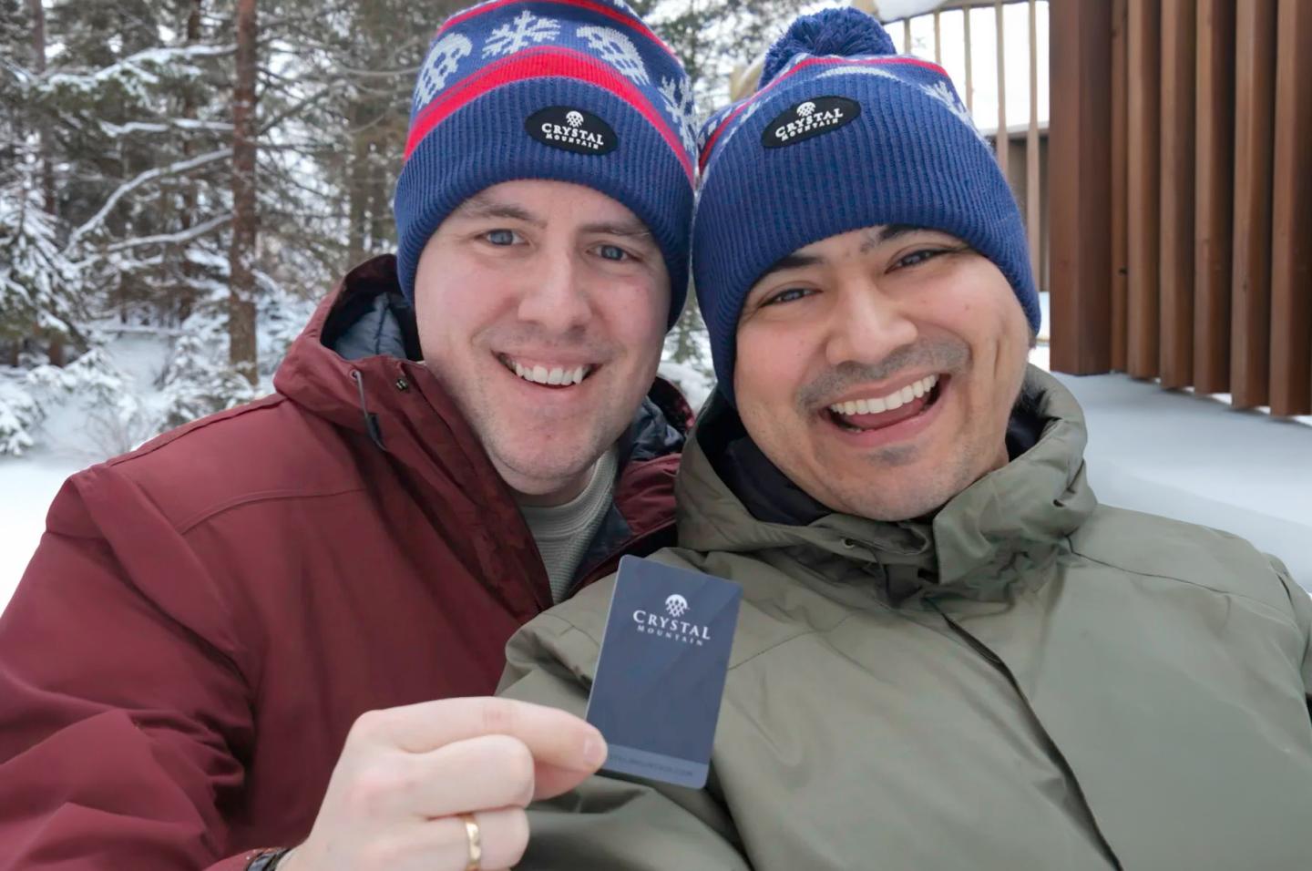 Two smiling people in winter clothing holding a key card, standing in a snowy forest.
