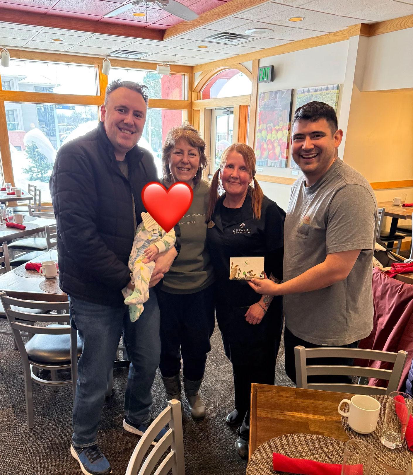 Four people smiling in a restaurant, one holding a baby with a heart covering its face.