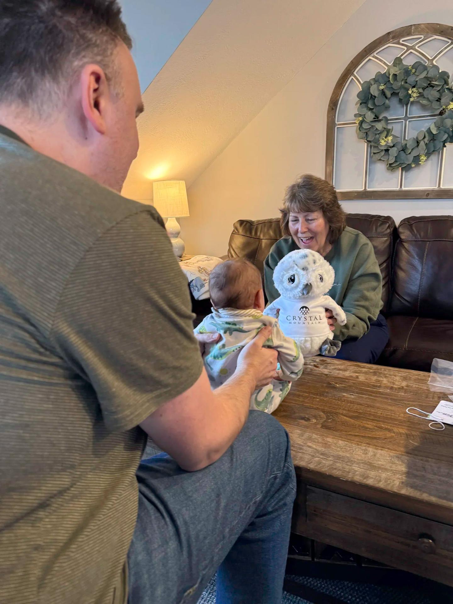 Man holding a baby, facing a smiling woman holding an owl toy in a cozy living room.