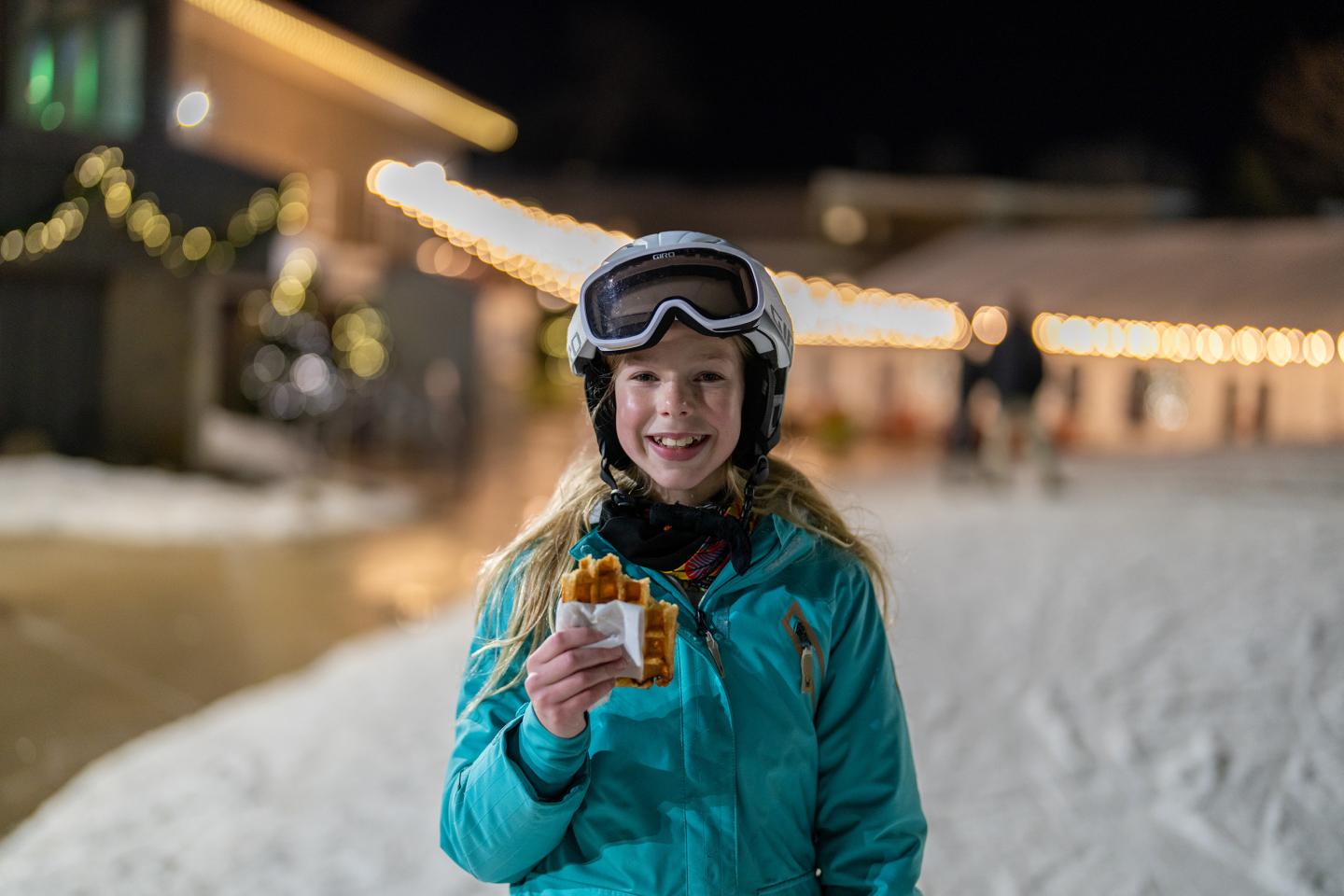 Girl in ski gear smiling with waffle, night setting.