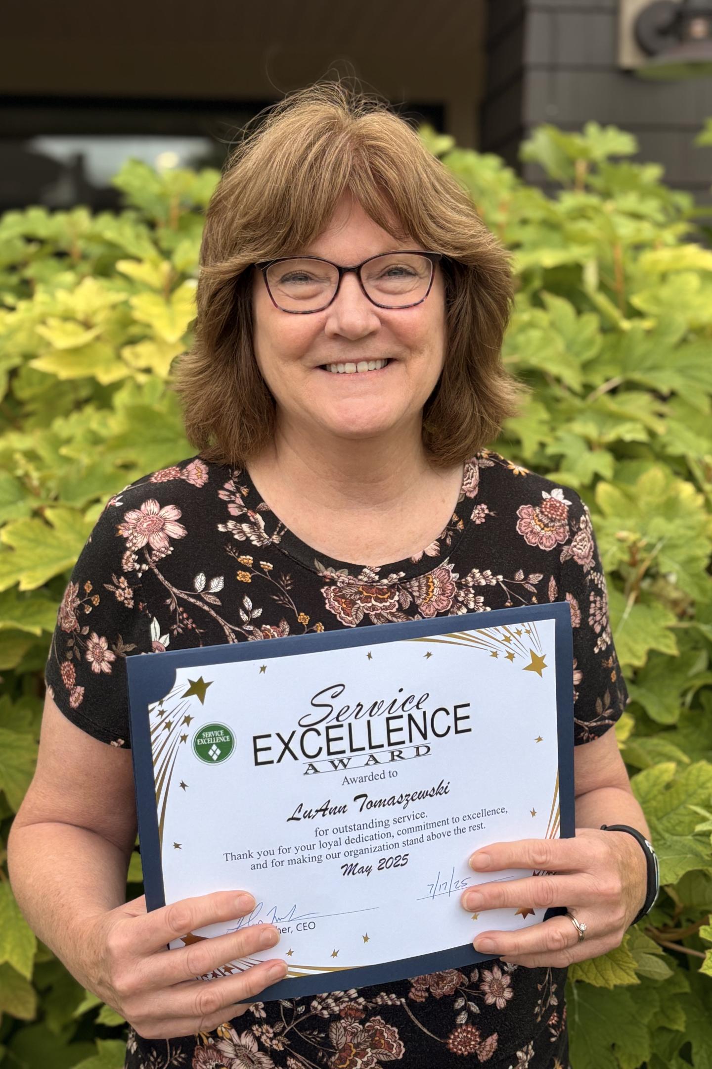 Smiling person holding an award certificate, standing in front of green leaves.