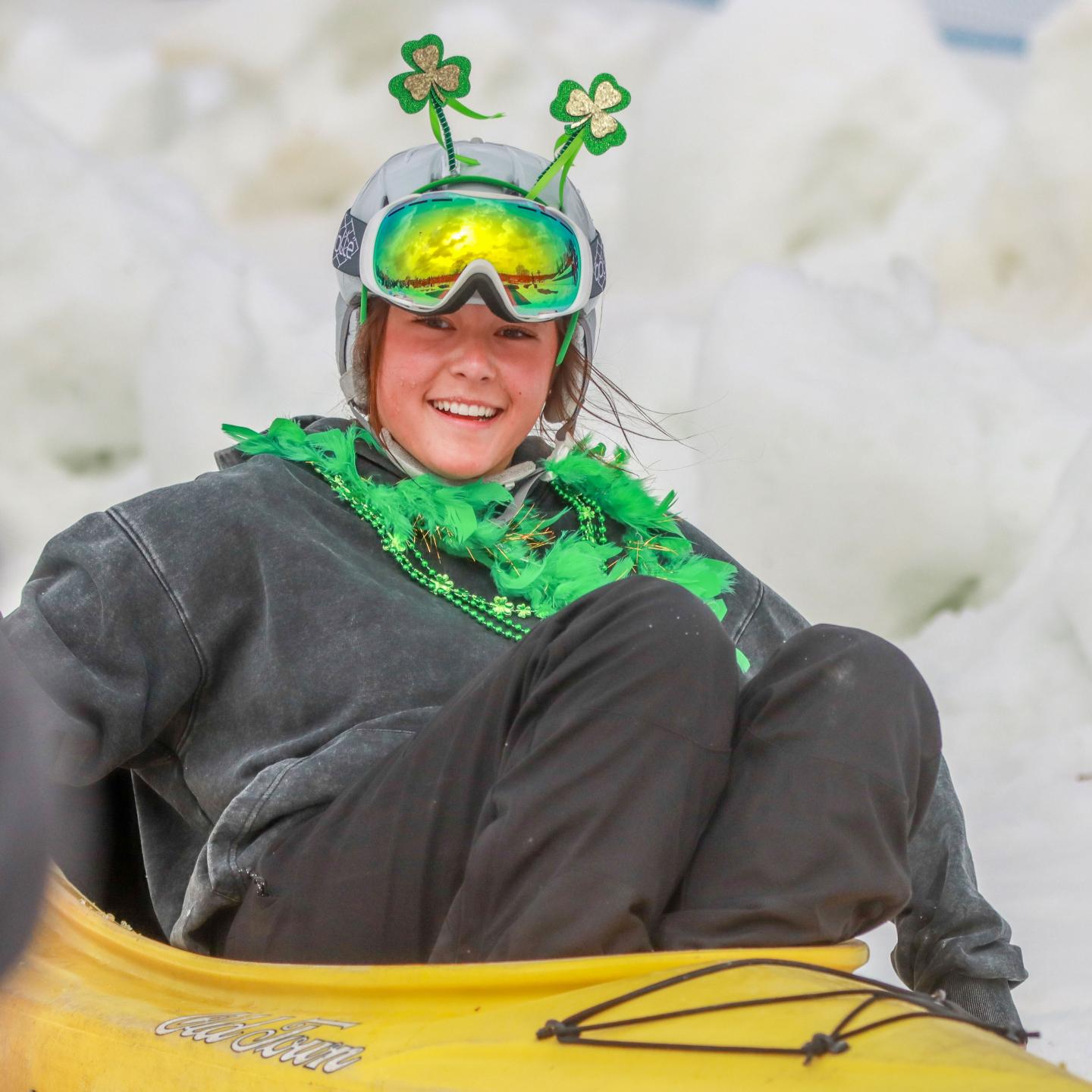 Person smiling in a yellow kayak, wearing goggles and shamrock accessories.