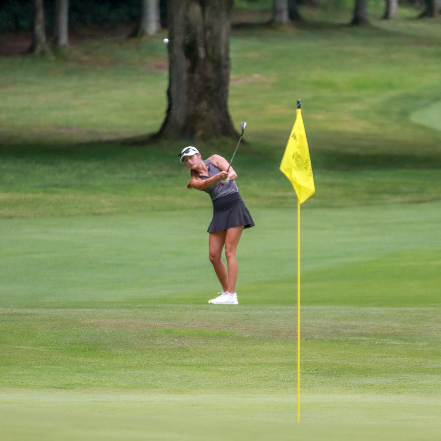 Golfer pitching towards the green near a flag on a lush golf course.