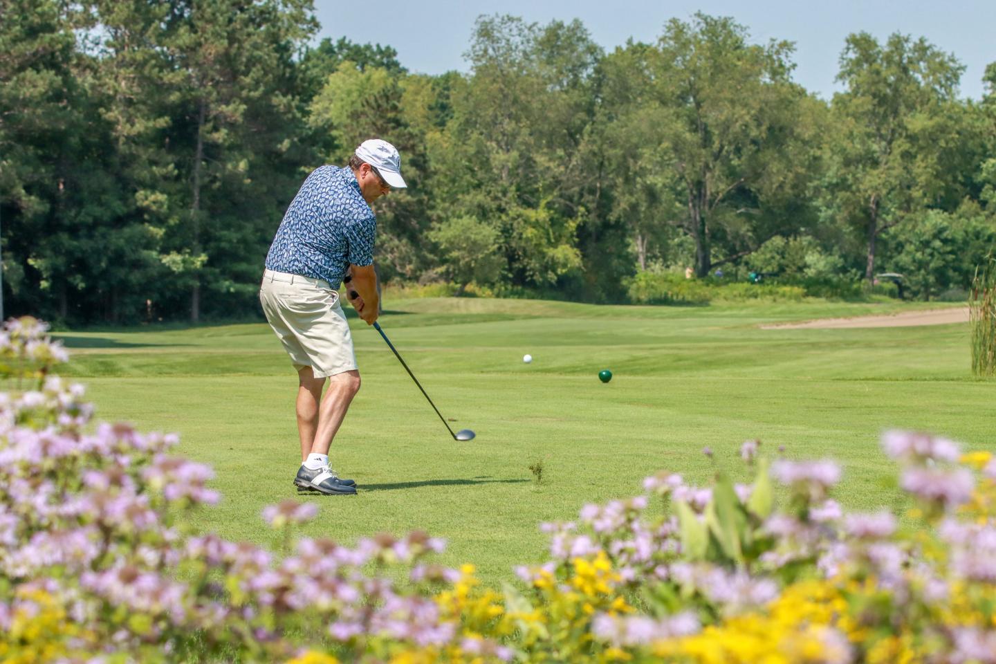 Golfer swings club on lush green course, surrounded by trees and colorful flowers.
