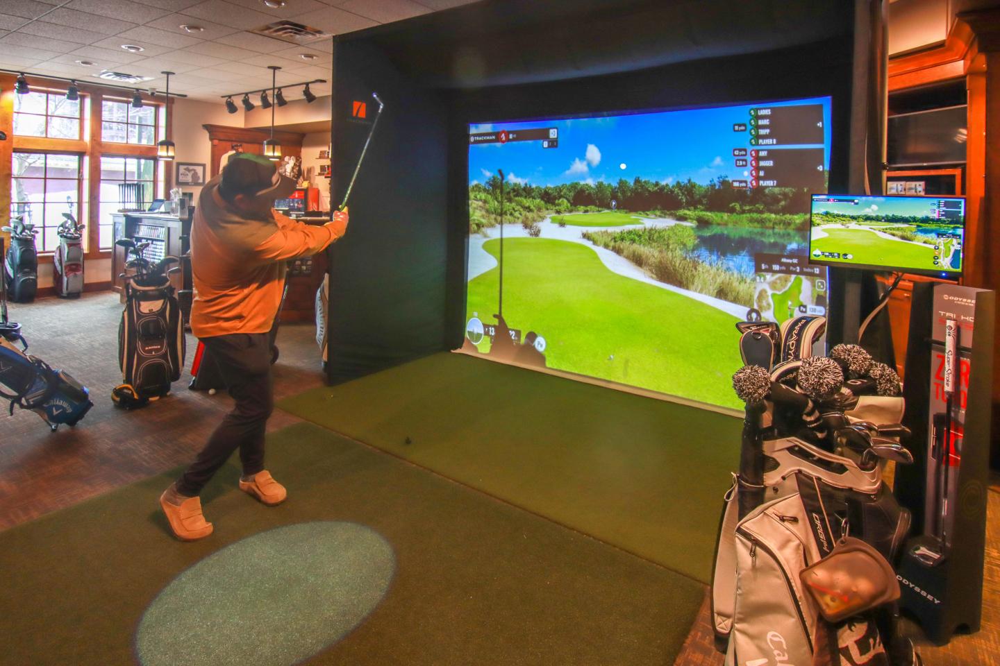 Golfer playing on an indoor simulator with golf bags nearby.