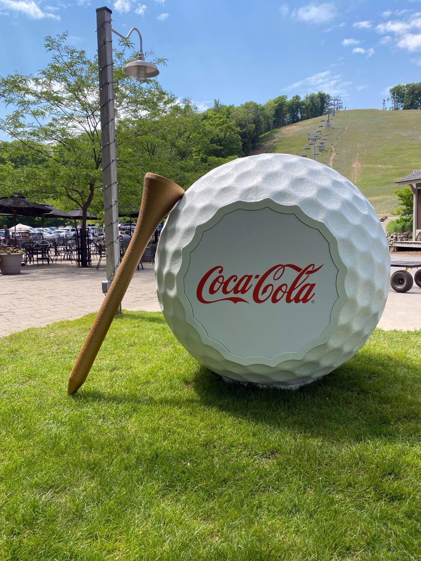 Giant golf ball with Coca-Cola logo on grass, next to a large wooden tee.