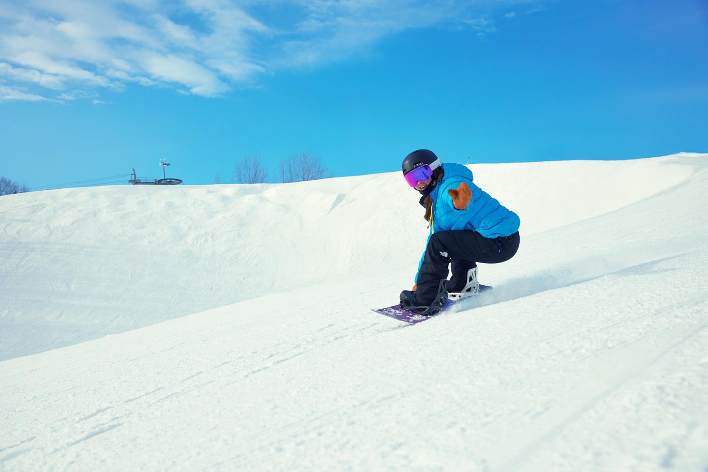 Snowboarder in blue jacket rides downhill on a sunny day.