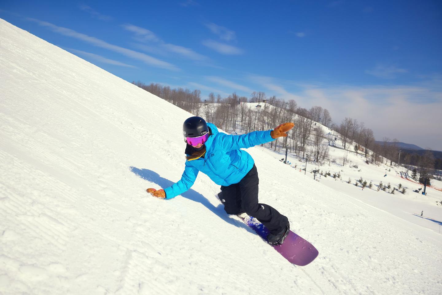 Snowboarder in blue jacket carving down snowy slope under clear blue sky.
