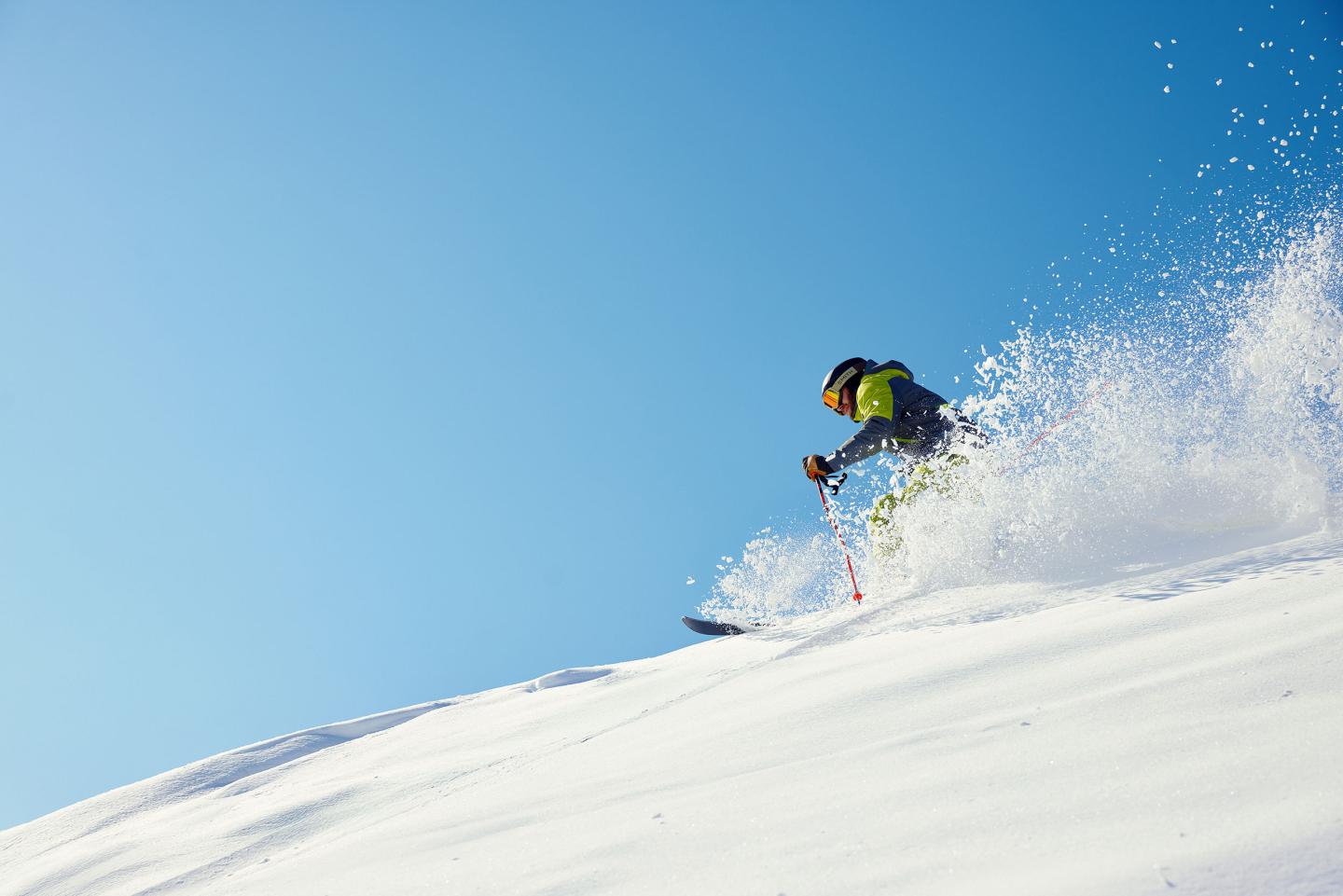 Skier in bright gear descending snowy slope against clear blue sky.