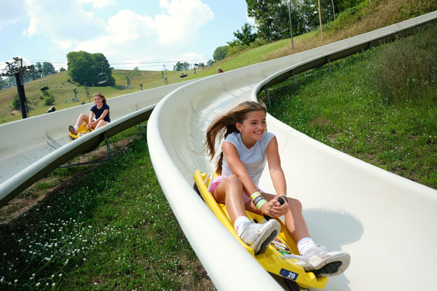Two people sliding down a summer alpine slide, smiling under sunny skies.