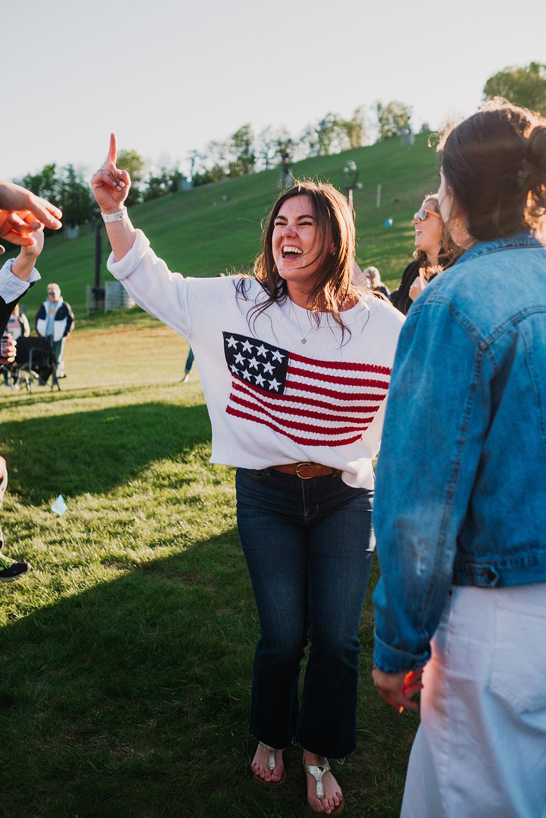 Woman in flag sweater smiling with friends, outdoors on sunny day.