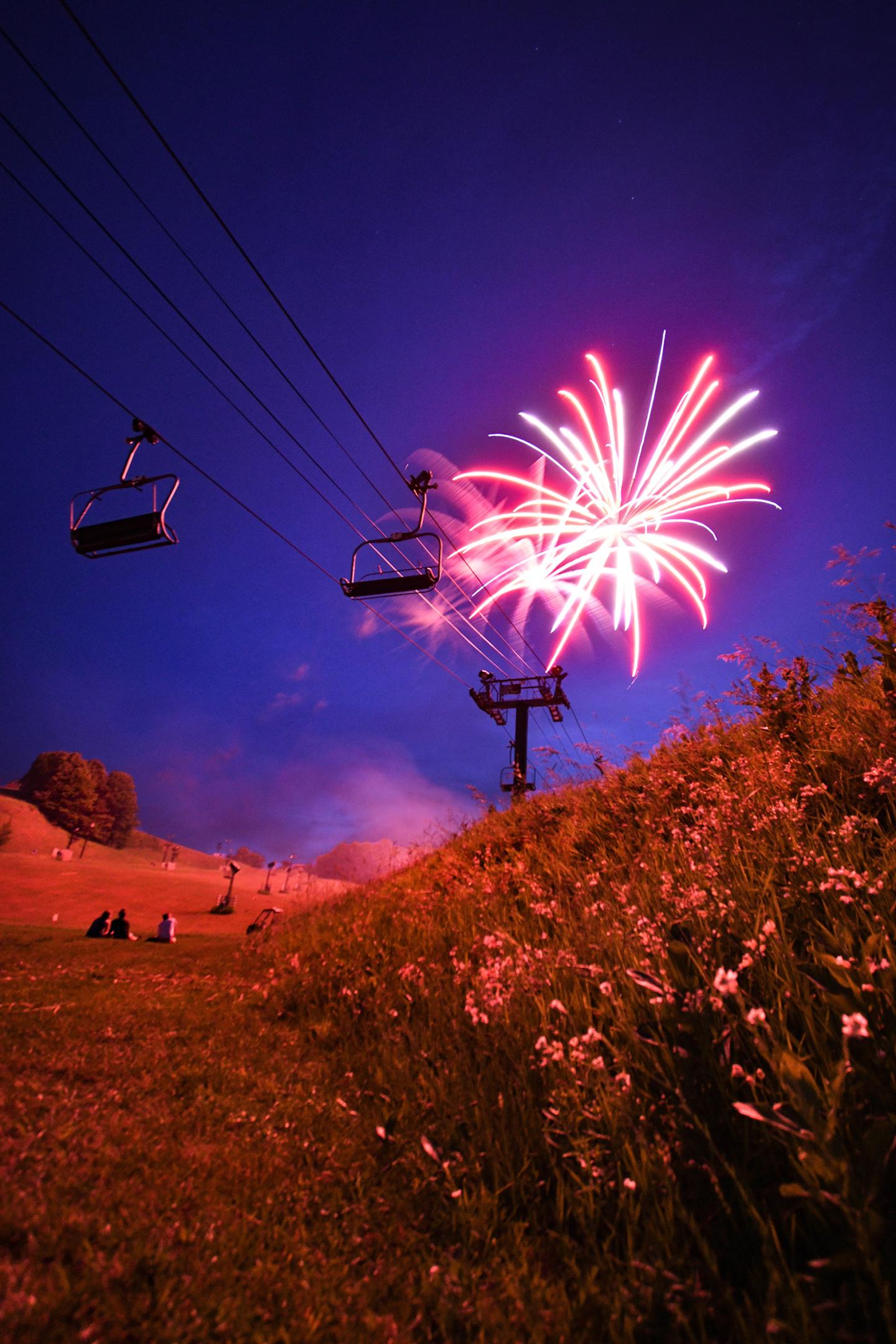 Fireworks light up a nighttime sky above a grassy hillside with a ski lift.