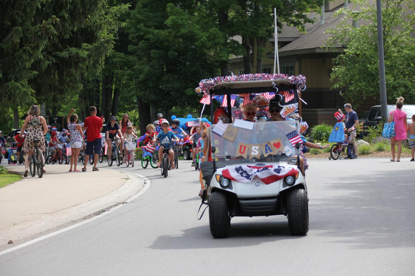 Parade with decorated golf cart, people walking, trees in background.